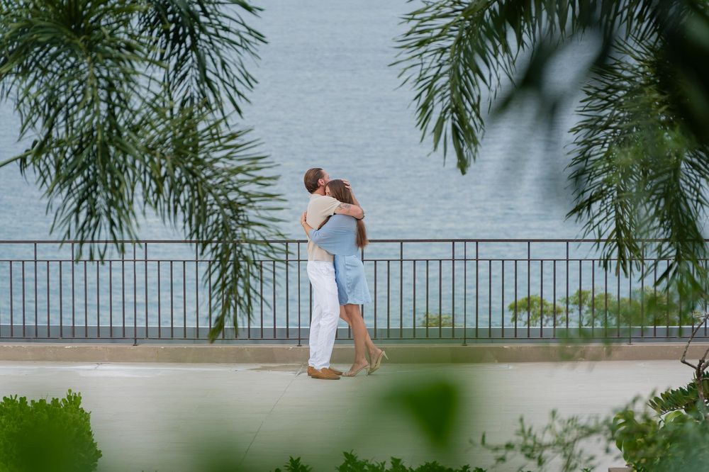 Couple embracing and kissing by a waterfront railing surrounded by tropical palm trees on a sunny day.