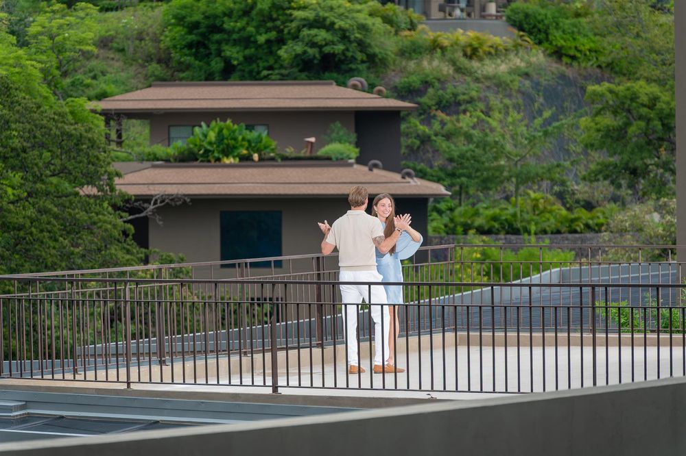 Couple standing on a railed overlook near a tropical brown building surrounded by lush green trees.