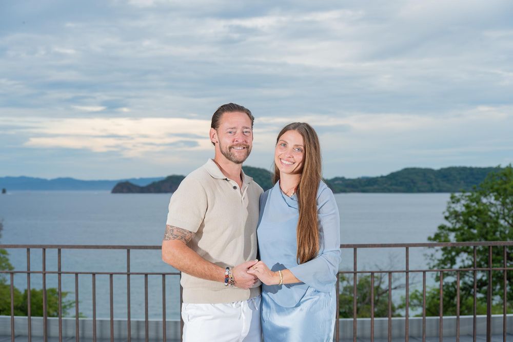 Smiling couple embracing on an outdoor terrace with scenic ocean and tropical island views in the background.