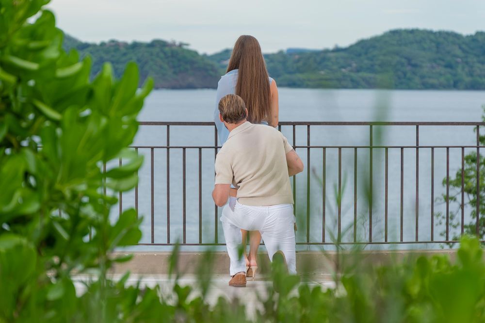 Man kneeling to propose to woman at scenic waterfront overlook with lush green hills in background.