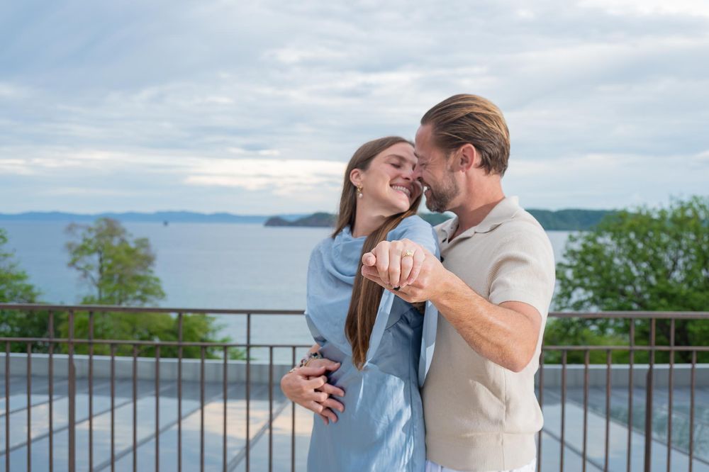 Couple embracing and smiling on a balcony overlooking the ocean on a cloudy day.