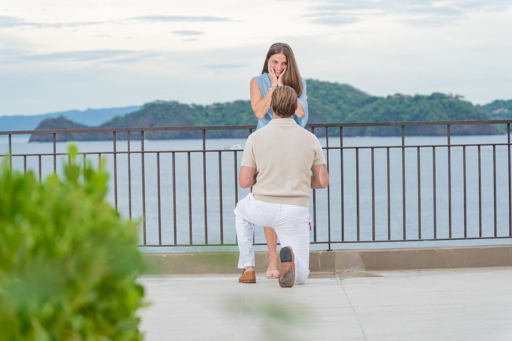 Surprised woman covers mouth as man proposes on one knee at beautiful ocean view terrace.