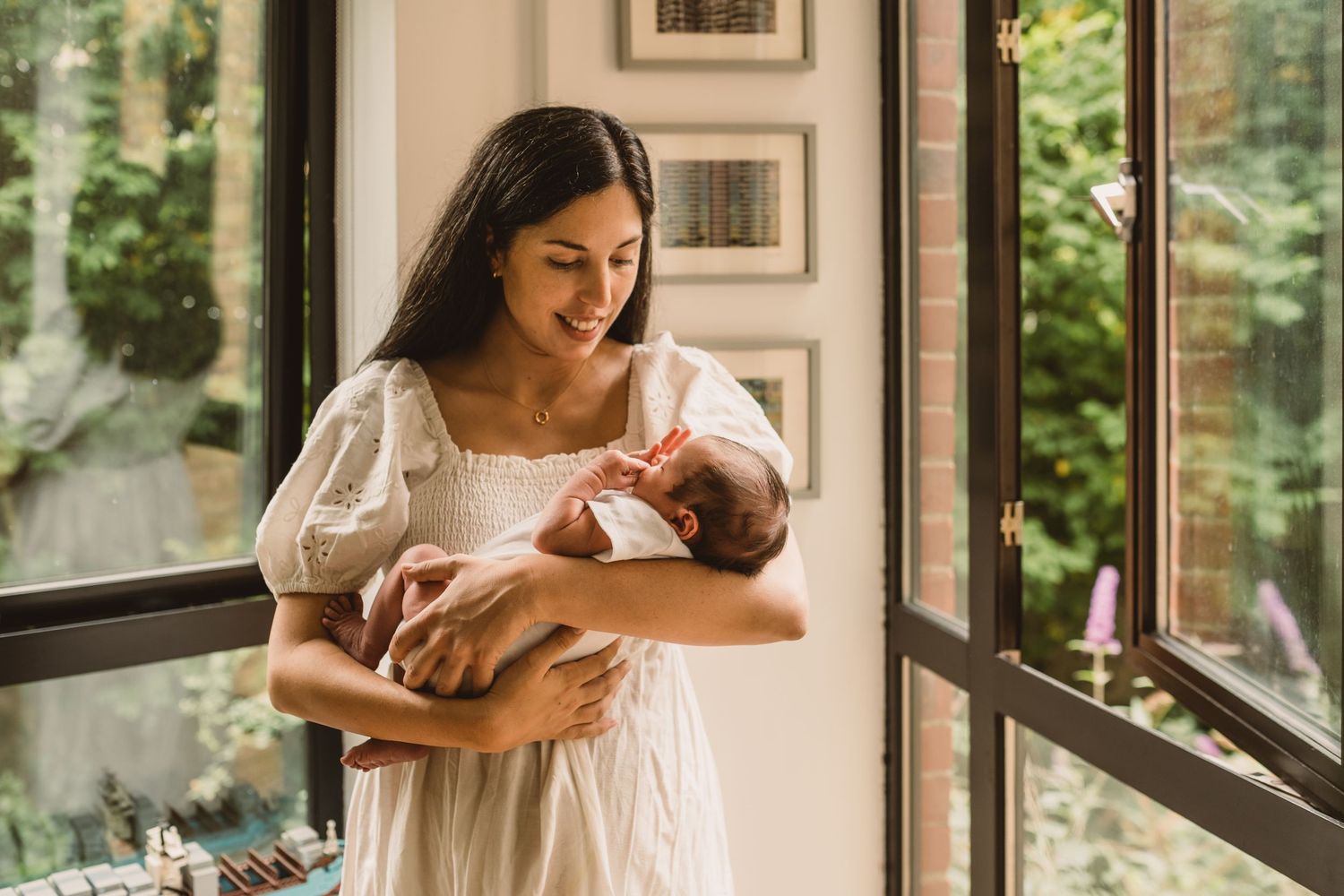 A smiling mother in a white dress tenderly holds her newborn baby near a sunlit window indoors.