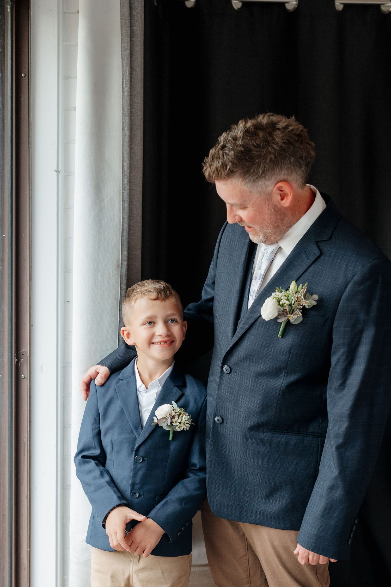 A man and young boy in matching navy suits with boutonnieres pose together near a window at a wedding.