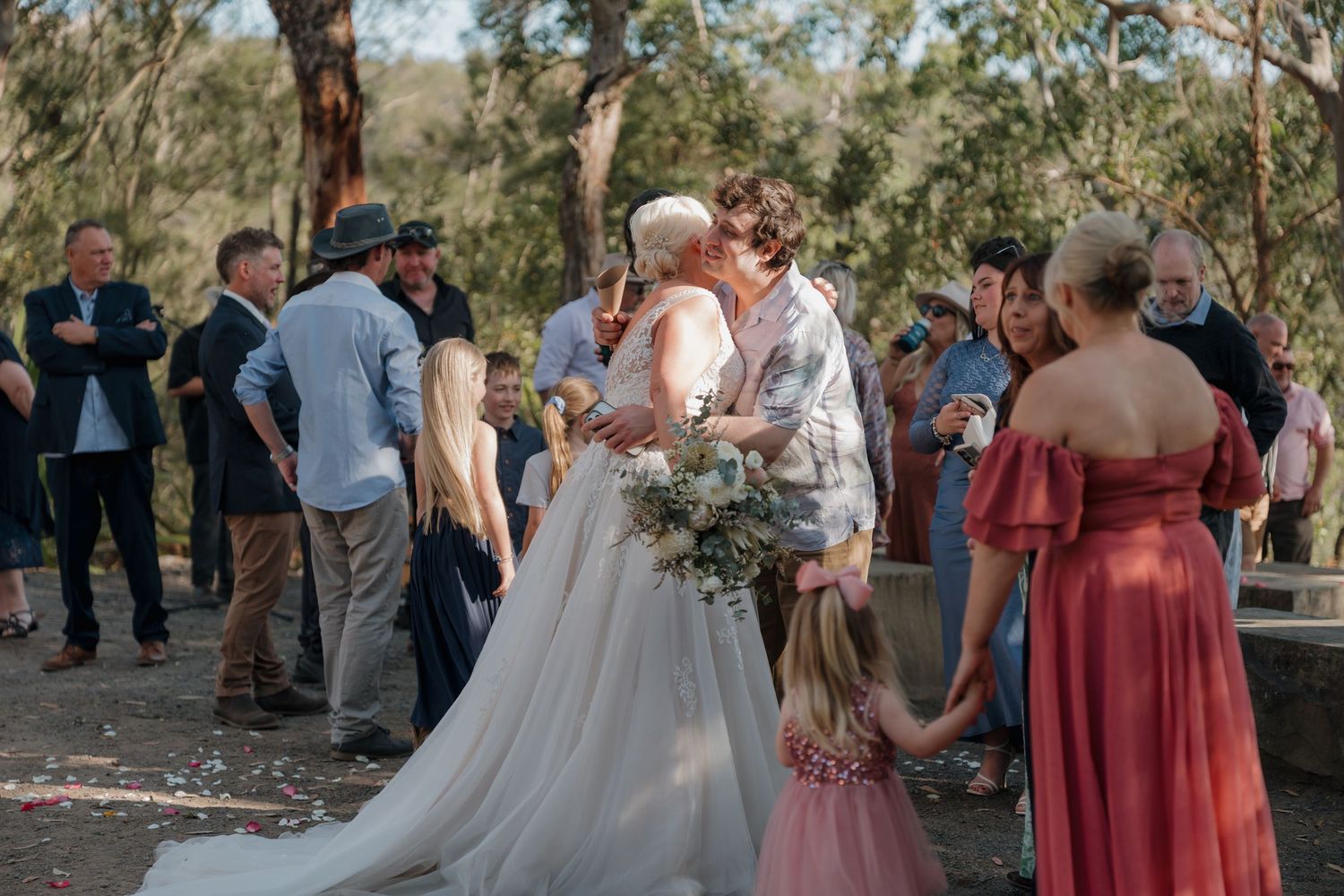 Bride and groom share a first kiss surrounded by wedding guests outdoors in a natural bush setting.