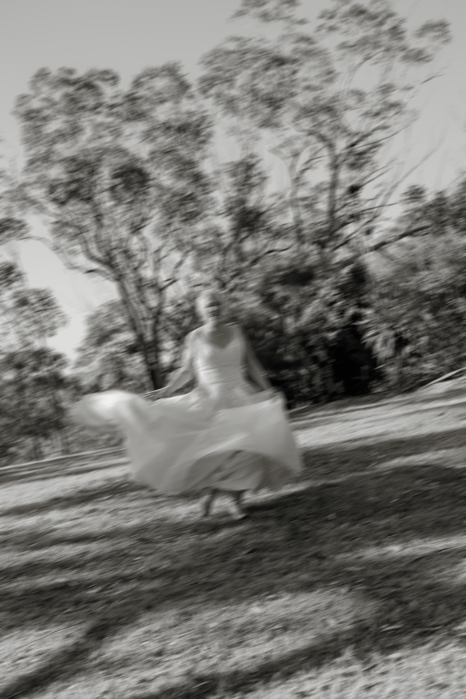 Blurred black and white photo of a bride twirling in a flowing wedding dress outdoors among trees.