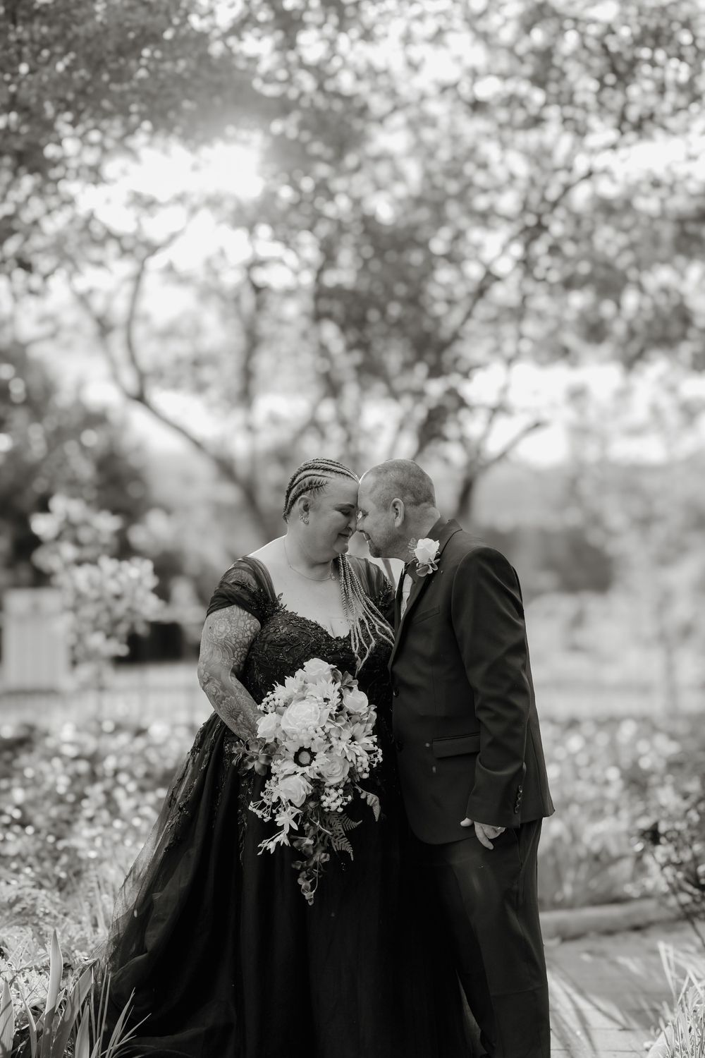 Black and white photo of a couple sharing an intimate moment, bride in dark gown holding bouquet, groom in suit, outdoors.