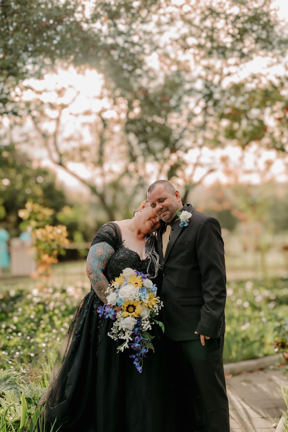 Couple in black wedding attire embracing outdoors, bride holding sunflower bouquet in blue and yellow.