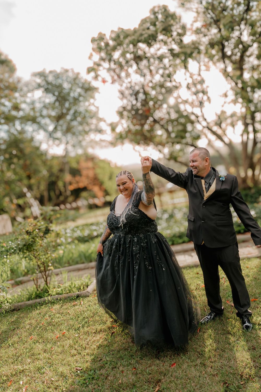 Couple dancing outdoors in matching black wedding attire, bride in black ball gown, groom in black suit, surrounded by lush greenery.
