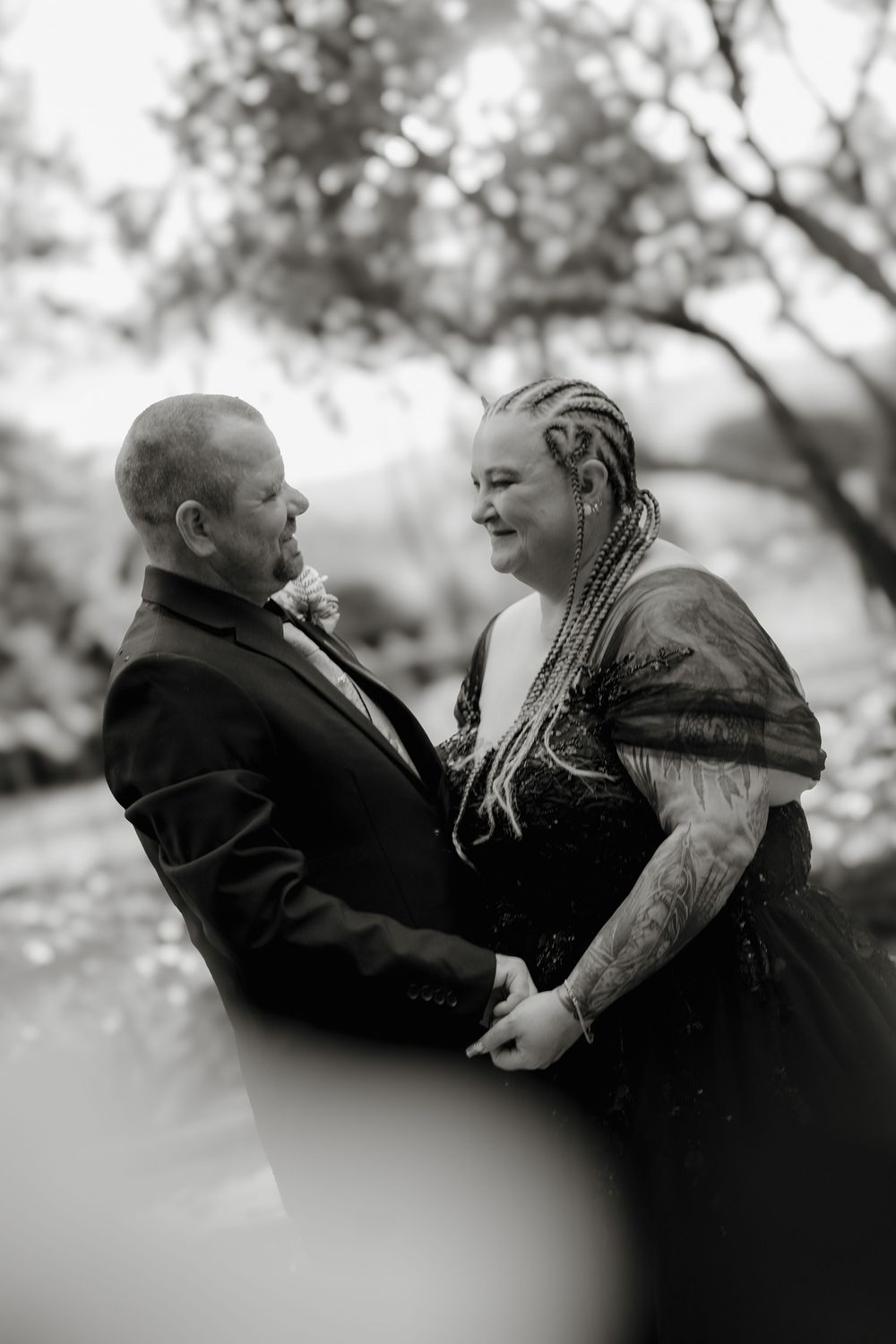 Black and white wedding photo of couple holding hands and gazing at each other outdoors, bride in black gown with braids.