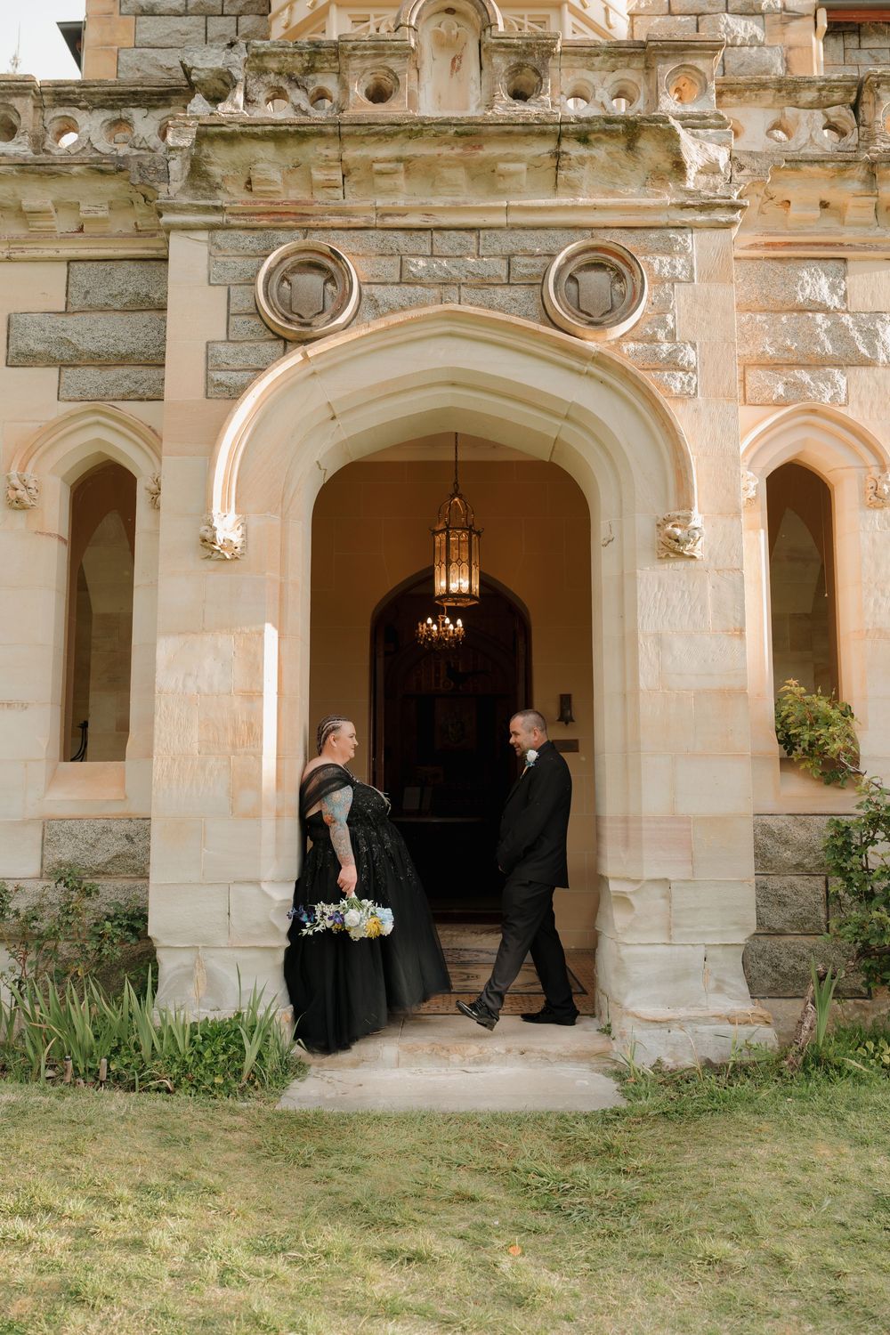 Couple in black wedding attire stand at ornate stone church entrance with arched doorway and chandelier.