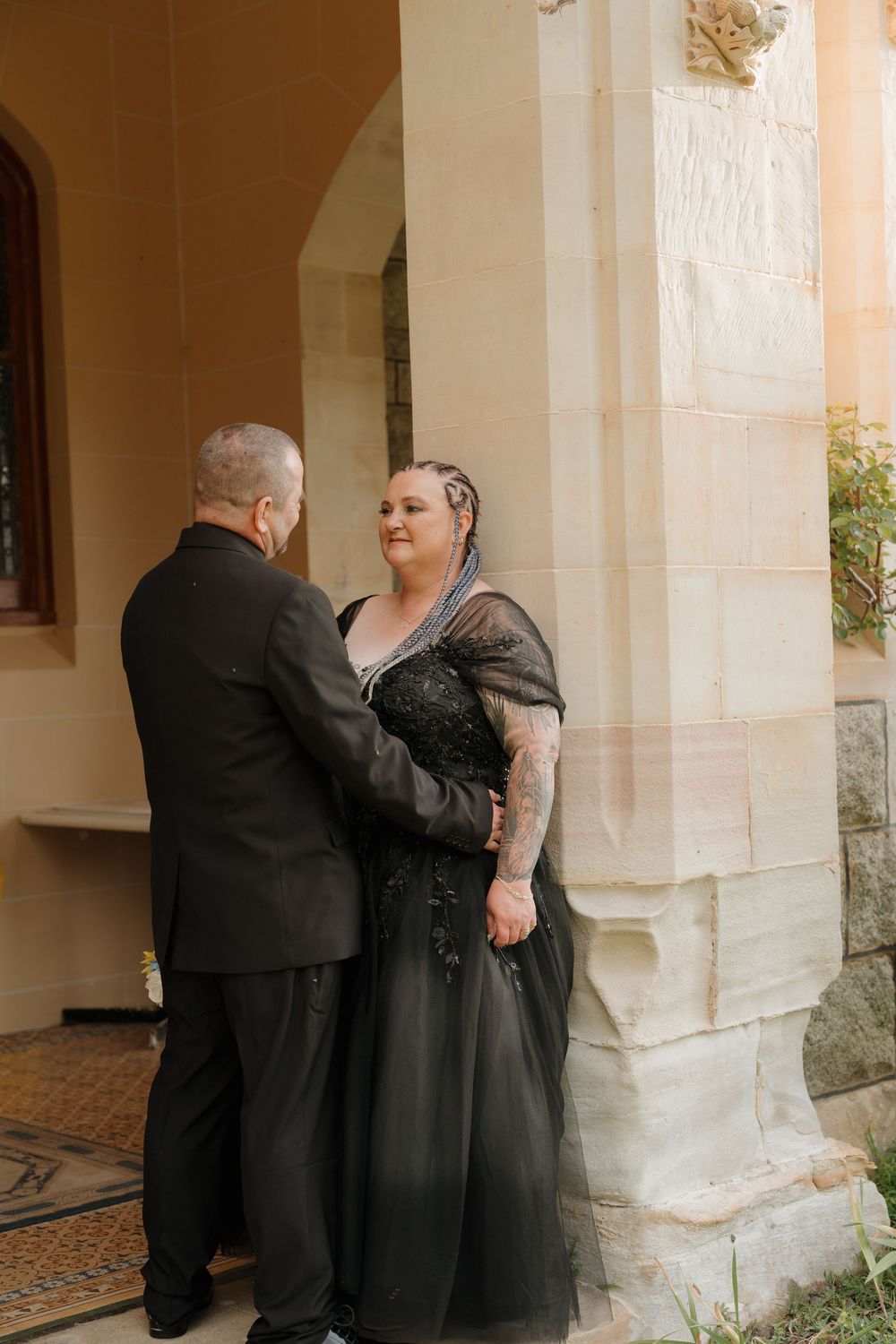 Couple in matching black wedding attire embracing outside a stone building, bride in black lace gown with tulle overlay.