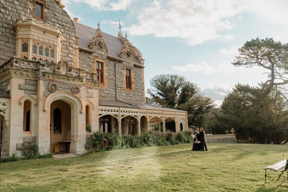 A couple in formal attire poses on the lawn of a grand Gothic stone mansion surrounded by lush gardens.