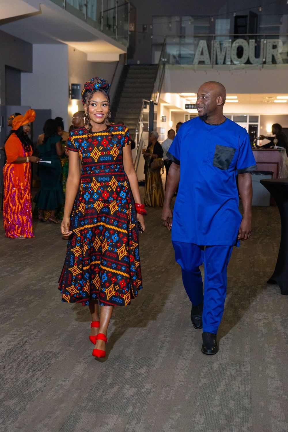 Couple in vibrant African print attire walking at an elegant event, woman in colorful dress, man in royal blue suit.