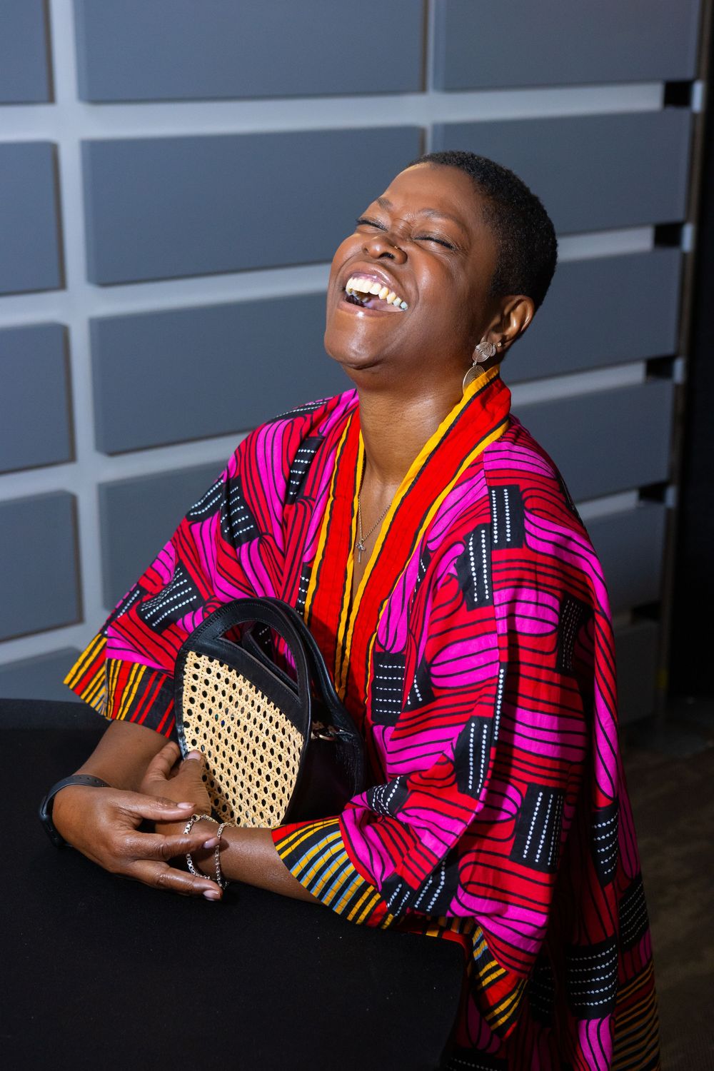 A joyful woman in a vibrant African print poncho laughs while holding a woven bag against a gray wall.