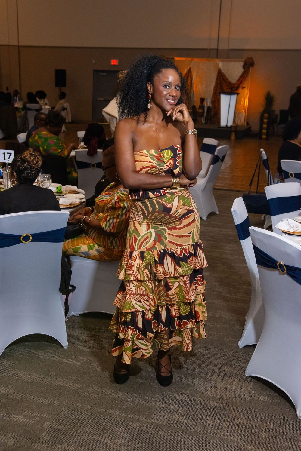 Woman in colorful African print strapless ruffle dress poses at elegant banquet hall event.