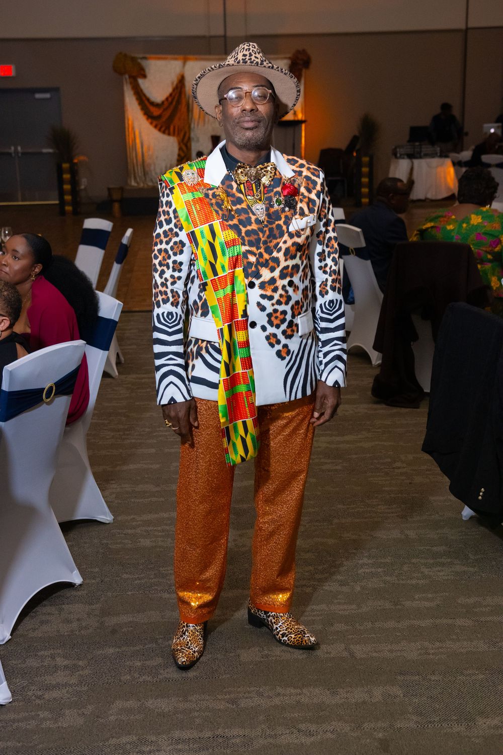 Man in vibrant African print blazer, leopard hat, orange sequin pants, and matching shoes at a formal event.