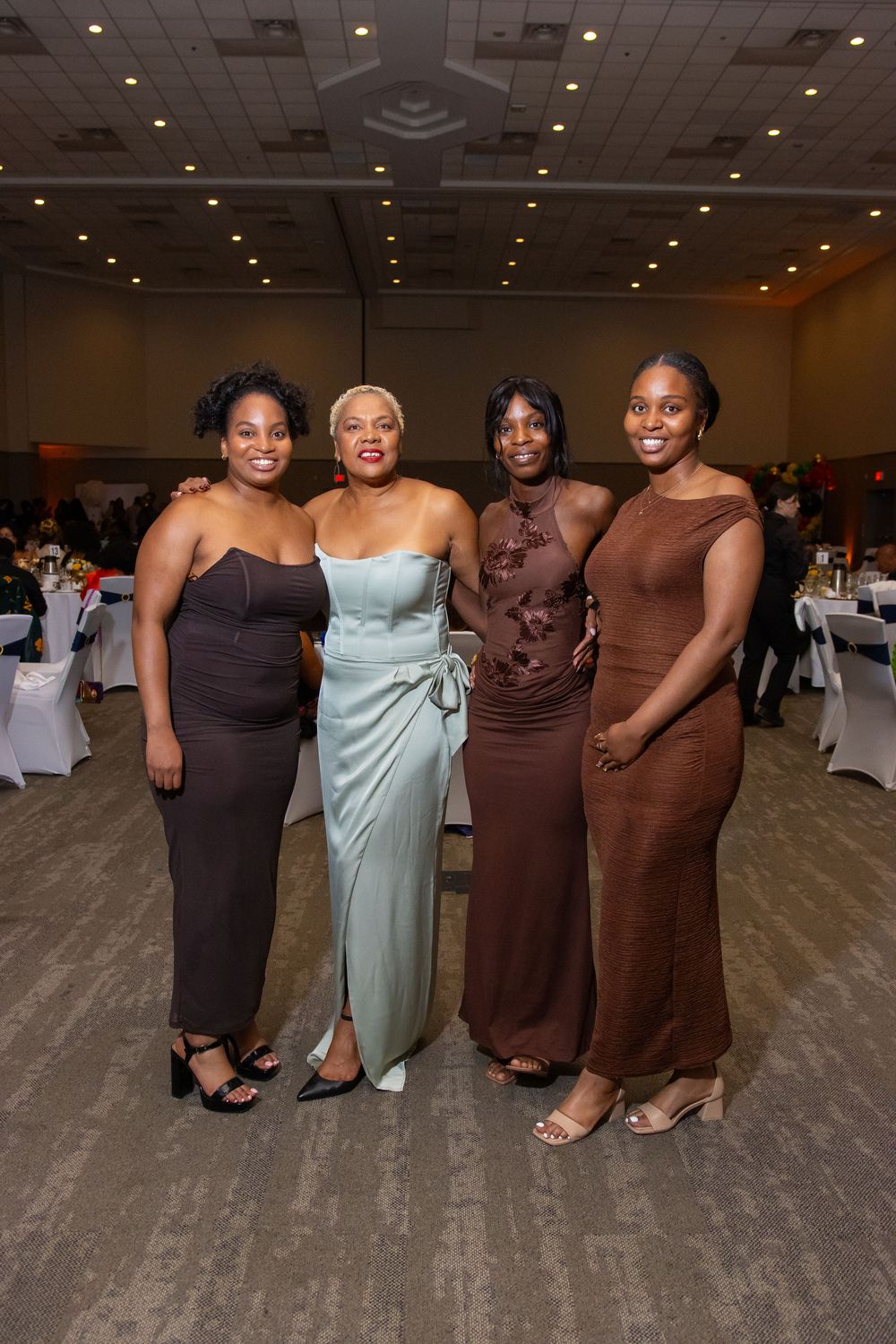 Four women in elegant formal dresses pose together smiling at a banquet hall event.