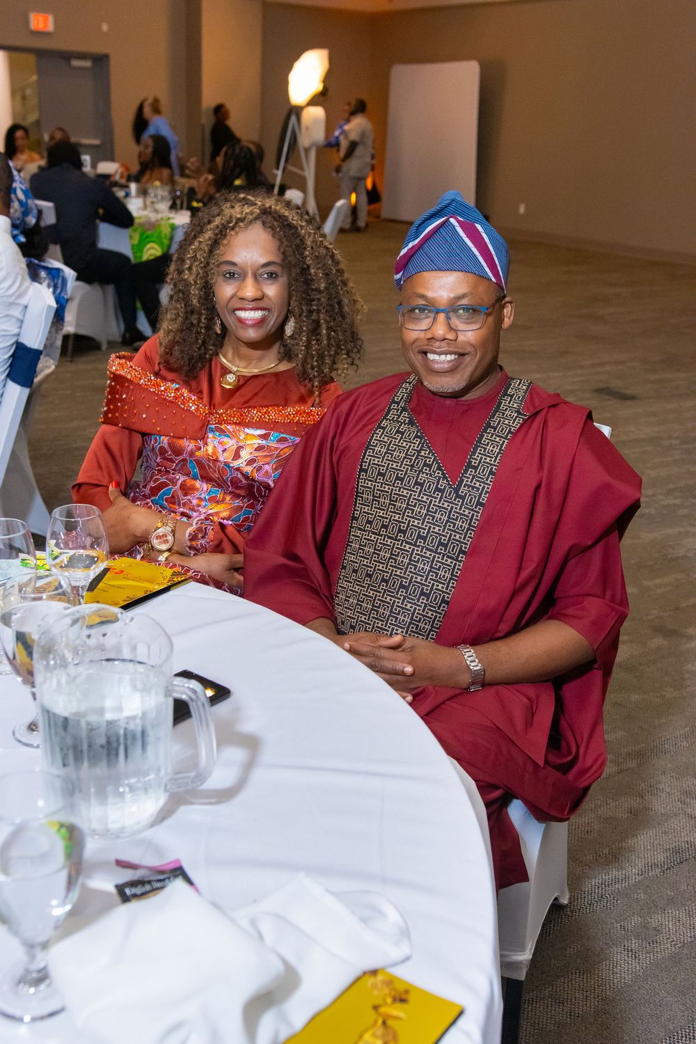 Two people in vibrant traditional African attire smile at a banquet table during a formal cultural event.