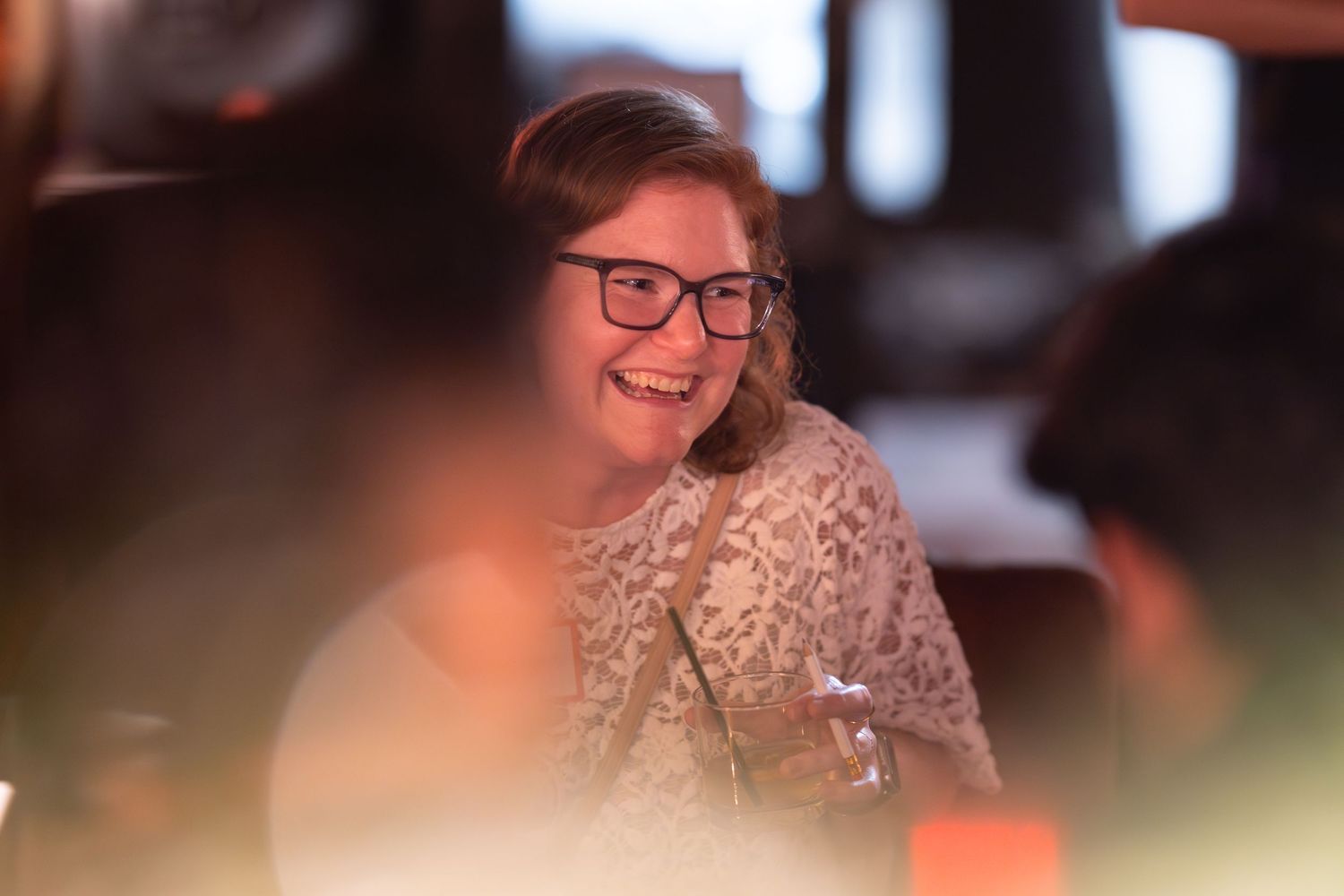 A smiling woman with glasses wearing a lace top enjoys a social gathering in a warmly lit venue.