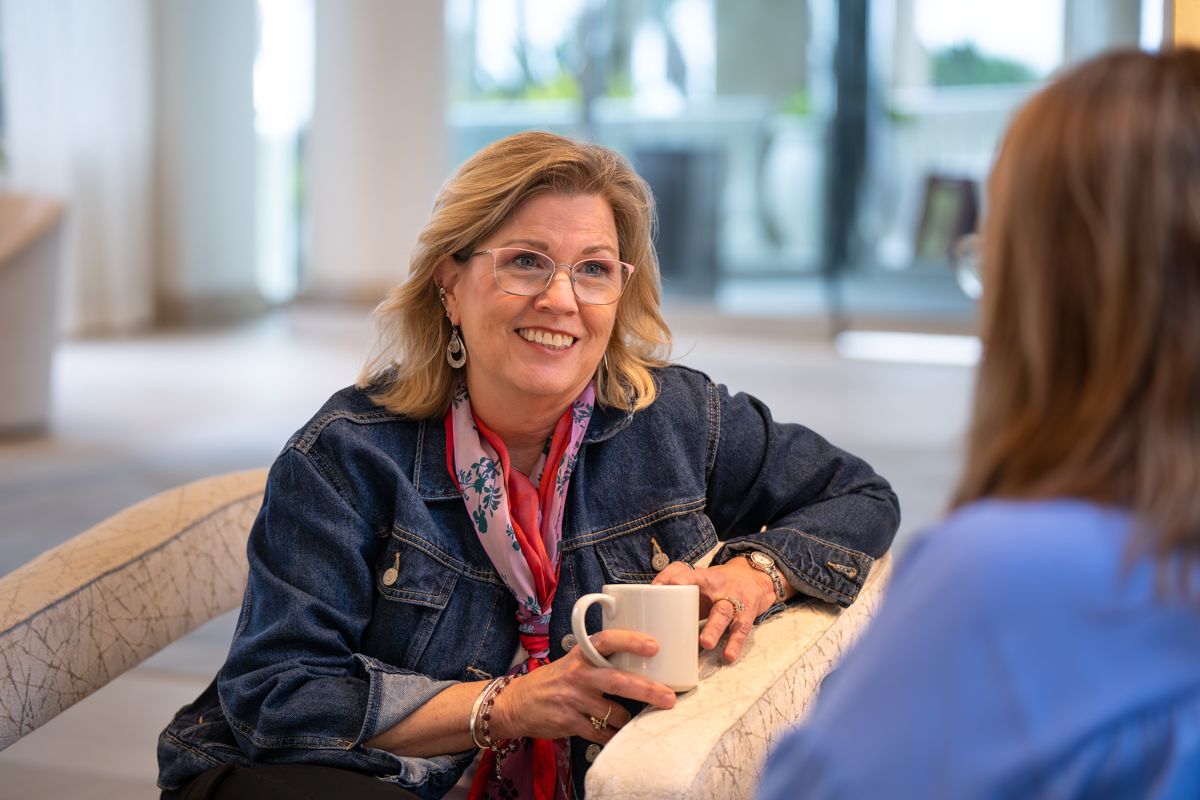 Smiling woman in denim jacket holding coffee mug, engaged in friendly conversation in a bright modern space.