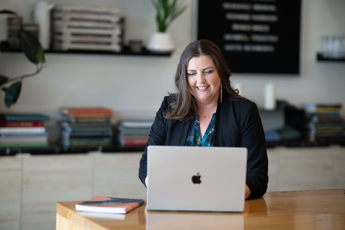 Woman smiling at Apple MacBook laptop at wooden desk in modern office with bookshelves and plants.