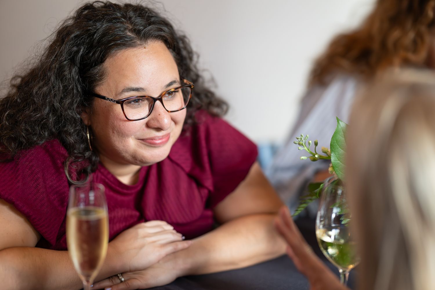 Woman with curly hair and glasses smiling at friend over champagne glasses at a social gathering.