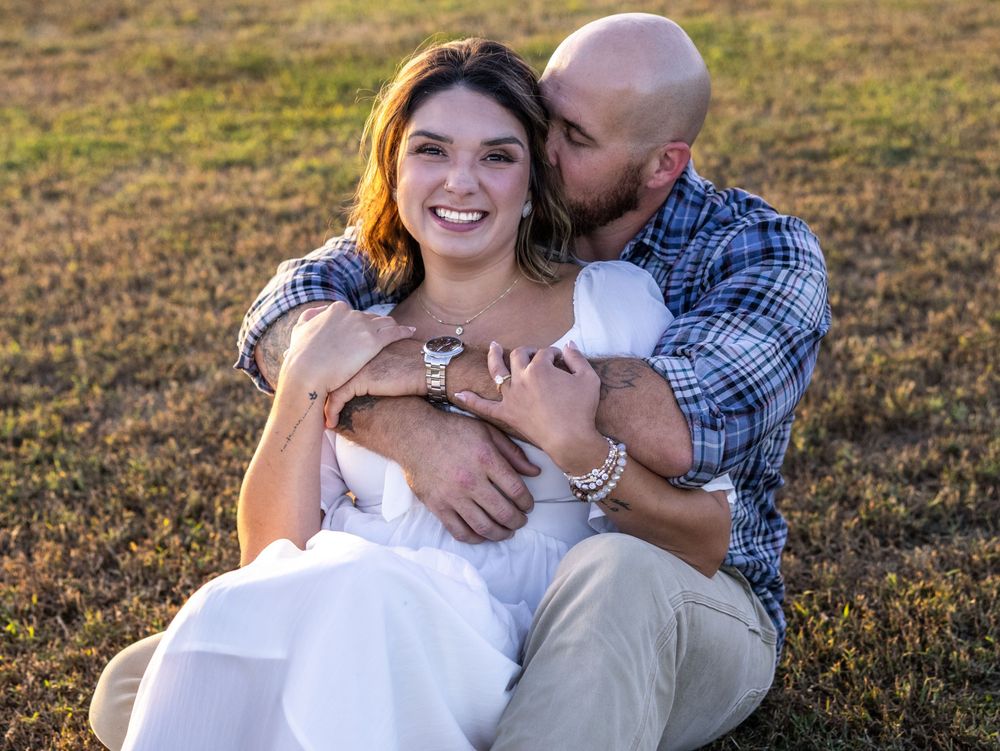 A happy couple sits on grass at golden hour, the man kissing the womans cheek as she smiles in a white dress.