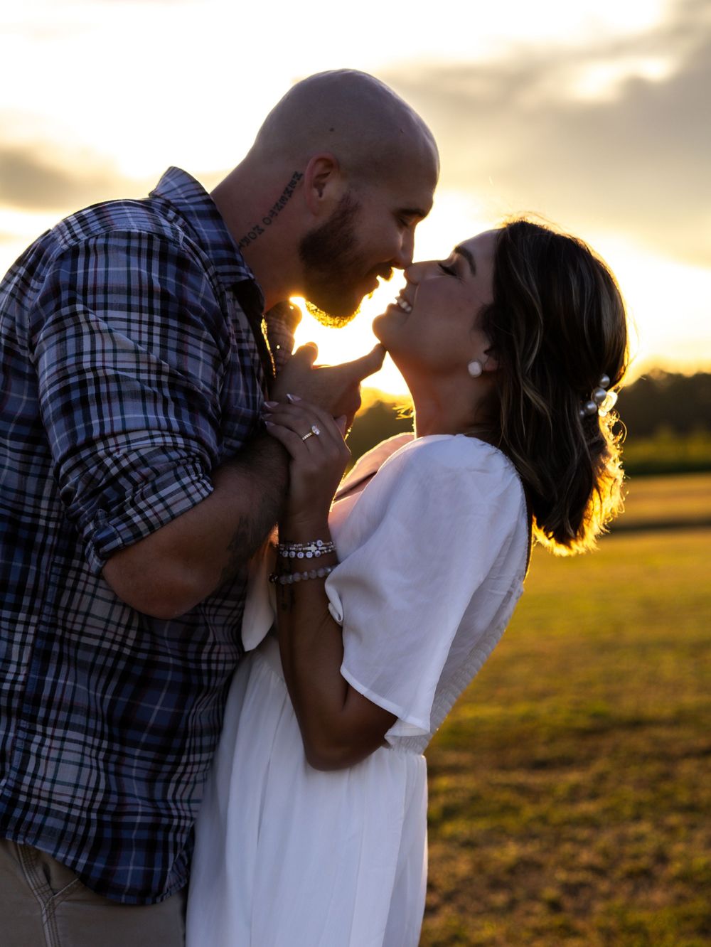 Couple sharing a romantic almost-kiss at golden sunset in an open field, man in plaid shirt, woman in white dress.