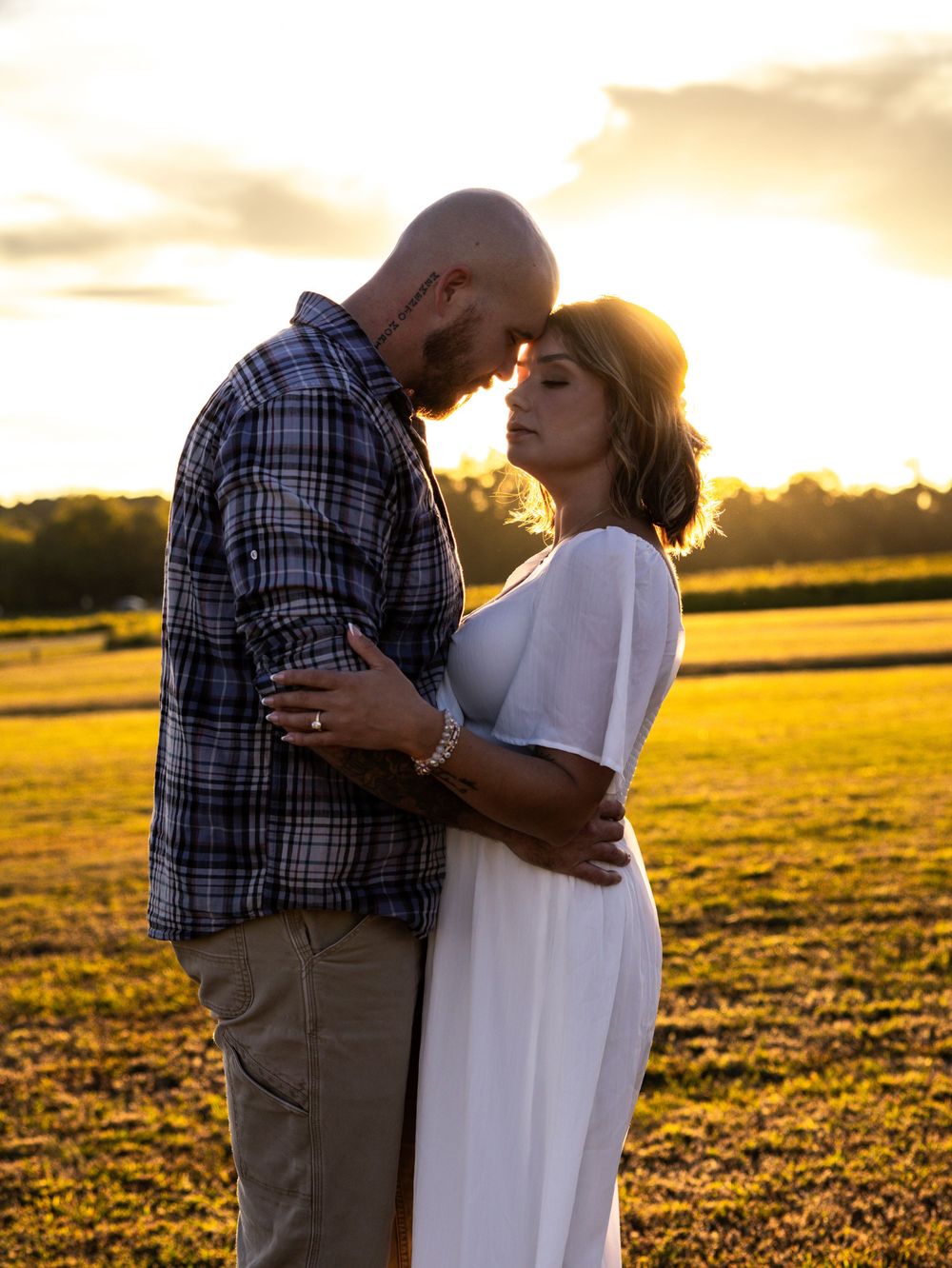 Couple embracing at golden sunset in open field, man in plaid shirt and woman in white dress touching foreheads romantically.
