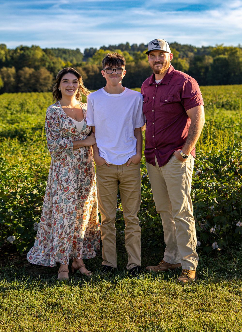 Family of three posing in a lush green vineyard on a sunny day, wearing casual outfits.