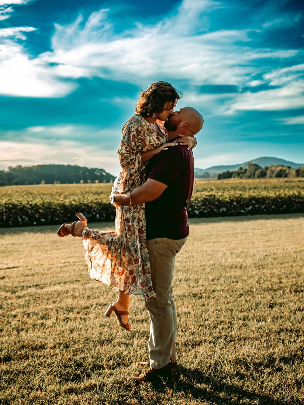 A couple shares a romantic kiss in a sunlit field, the man lifting the woman in a floral dress against a blue sky.