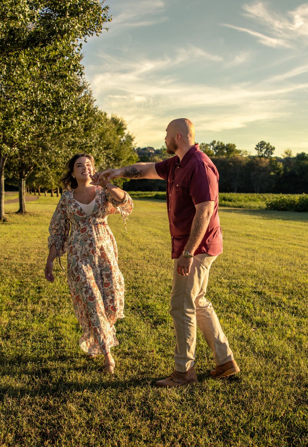 A couple dances together in a sunlit green field, the woman in a floral dress and the man in a burgundy shirt.