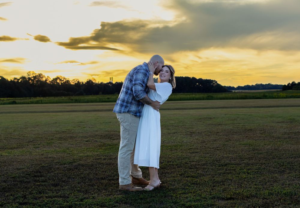 Couple sharing a romantic kiss at sunset in an open green field with dramatic cloudy sky backdrop.