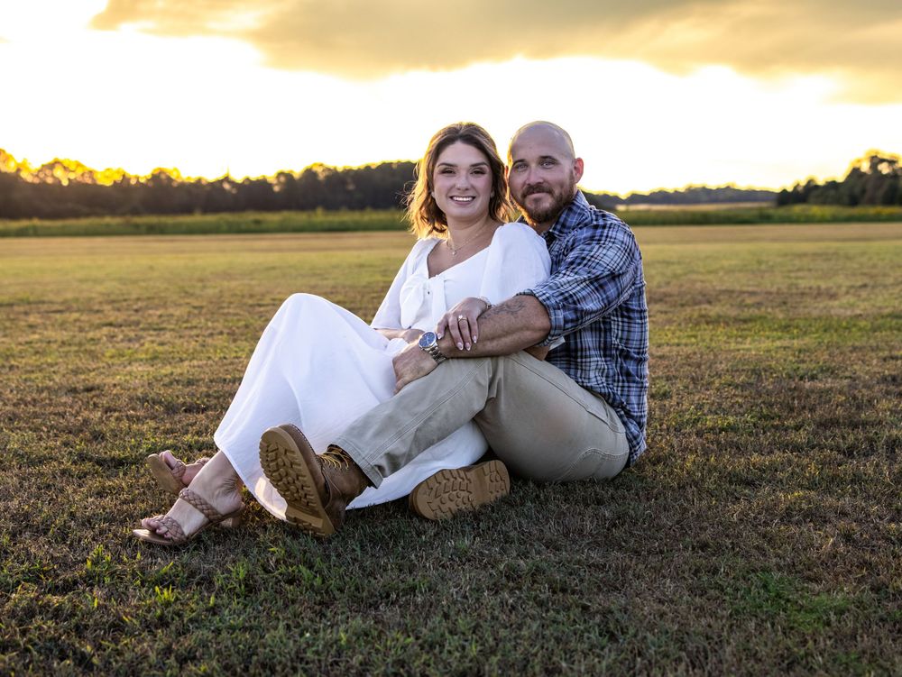Couple sitting together on a grassy field at golden hour sunset, smiling warmly.