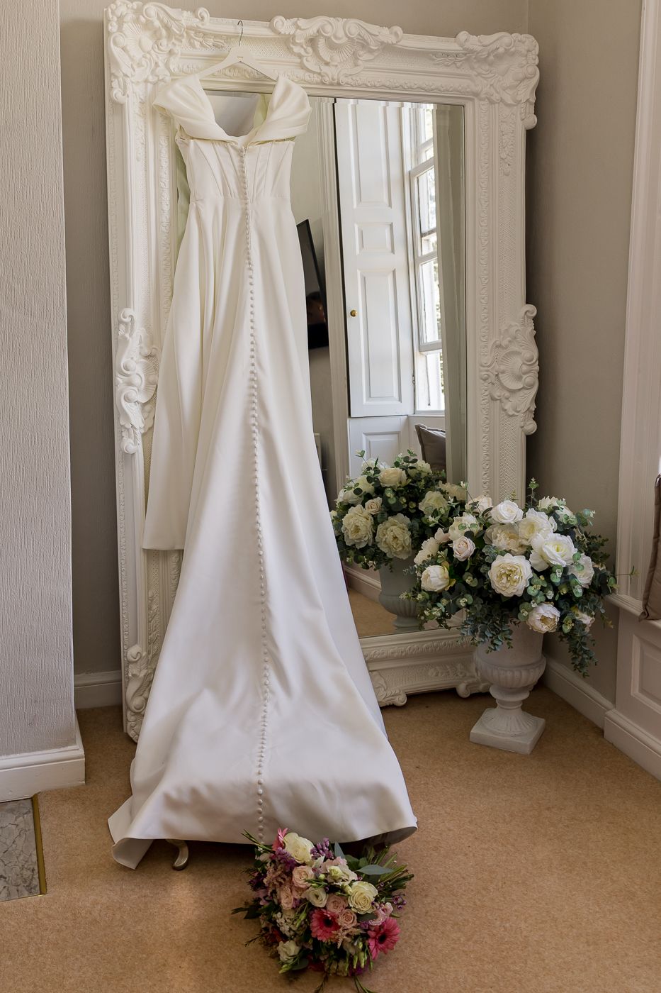 Elegant white satin wedding dress hanging on an ornate white mirror with floral bouquets on the floor.