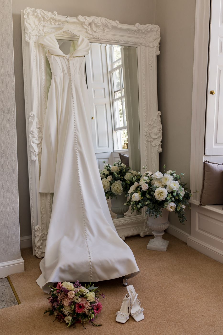 Elegant white satin wedding dress hanging on ornate mirror with bridal bouquet and heels on floor.