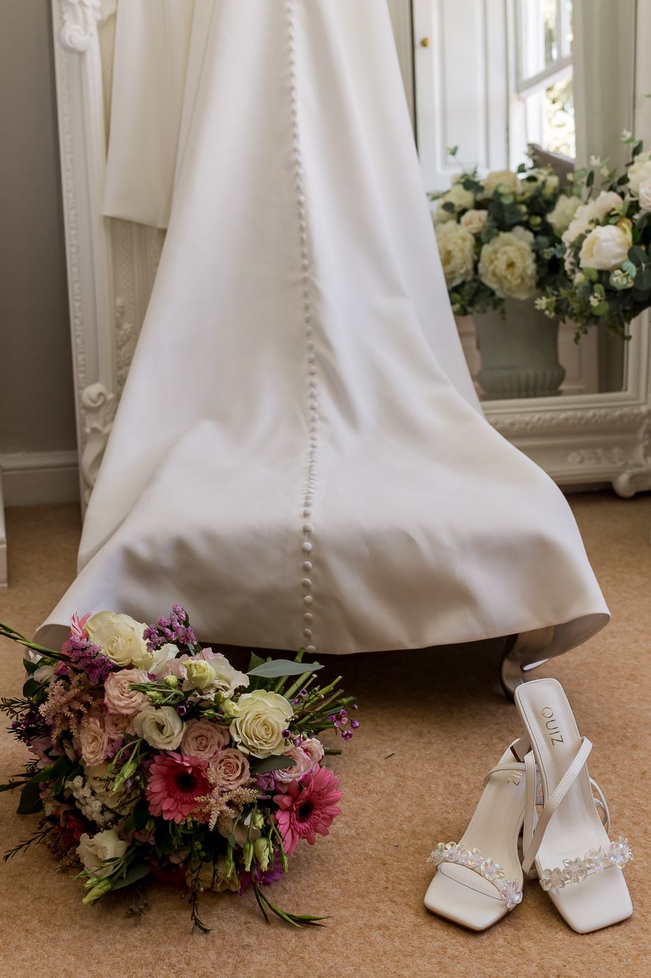 Elegant wedding dress train displayed beside bridal bouquet of pink roses and white heels on carpeted floor.