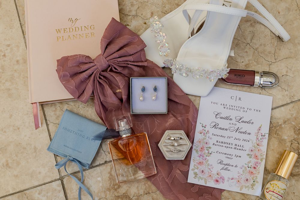 Elegant wedding flat lay with floral invitation, diamond rings, pearl earrings, perfume, bridal heels, and a pink bow on tile.