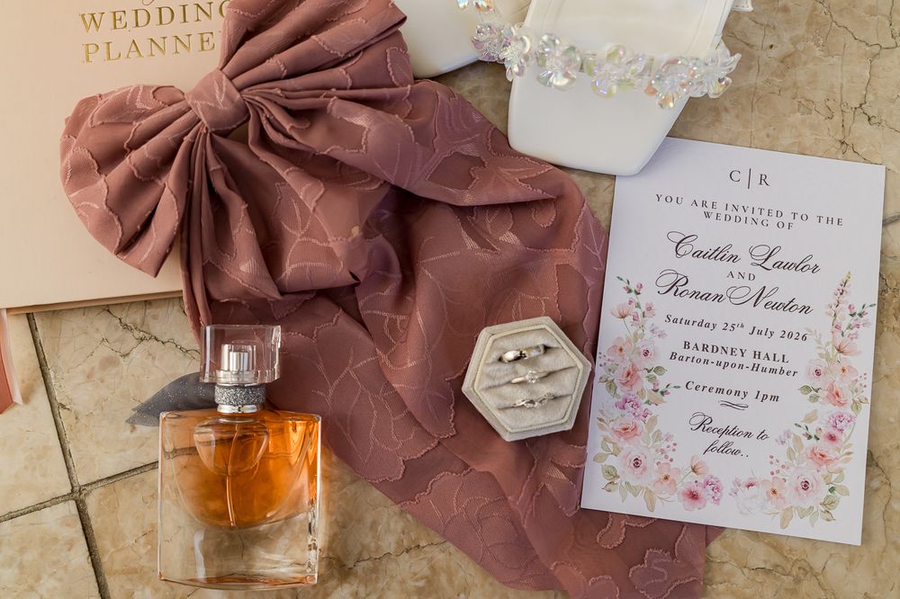 Close-up wedding flat lay featuring floral invitation, engagement rings, perfume bottle, and dusty rose bow accent.