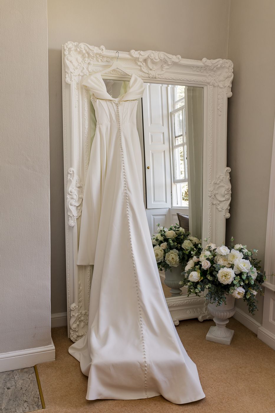 Elegant white satin wedding dress hanging on ornate white mirror with floral bouquet arrangement in a bridal suite.