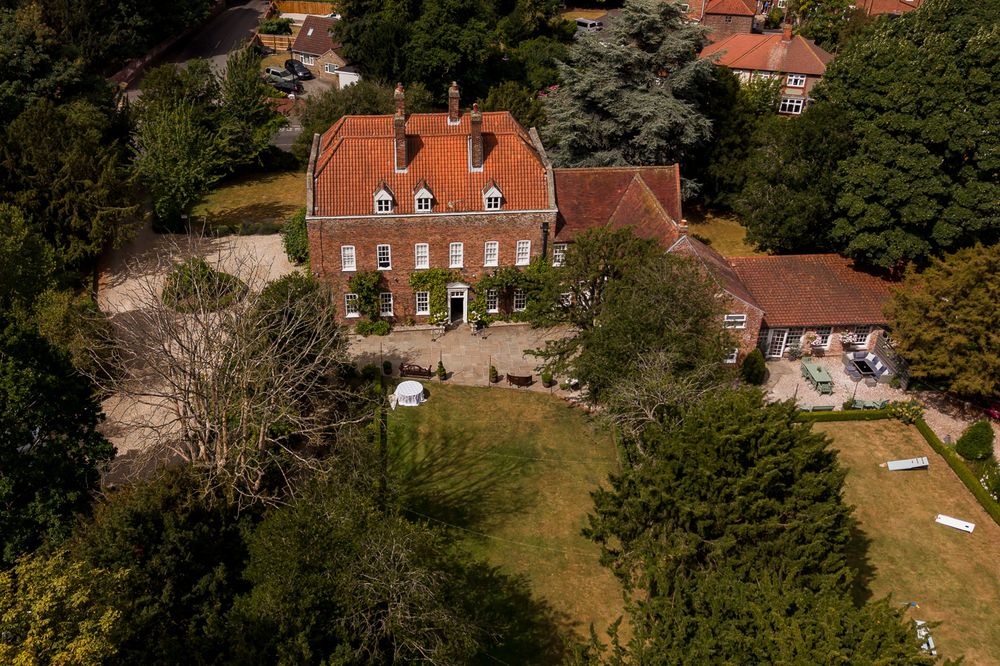 Aerial view of a grand red-brick Georgian country house with terracotta roof, manicured lawn, and mature trees.