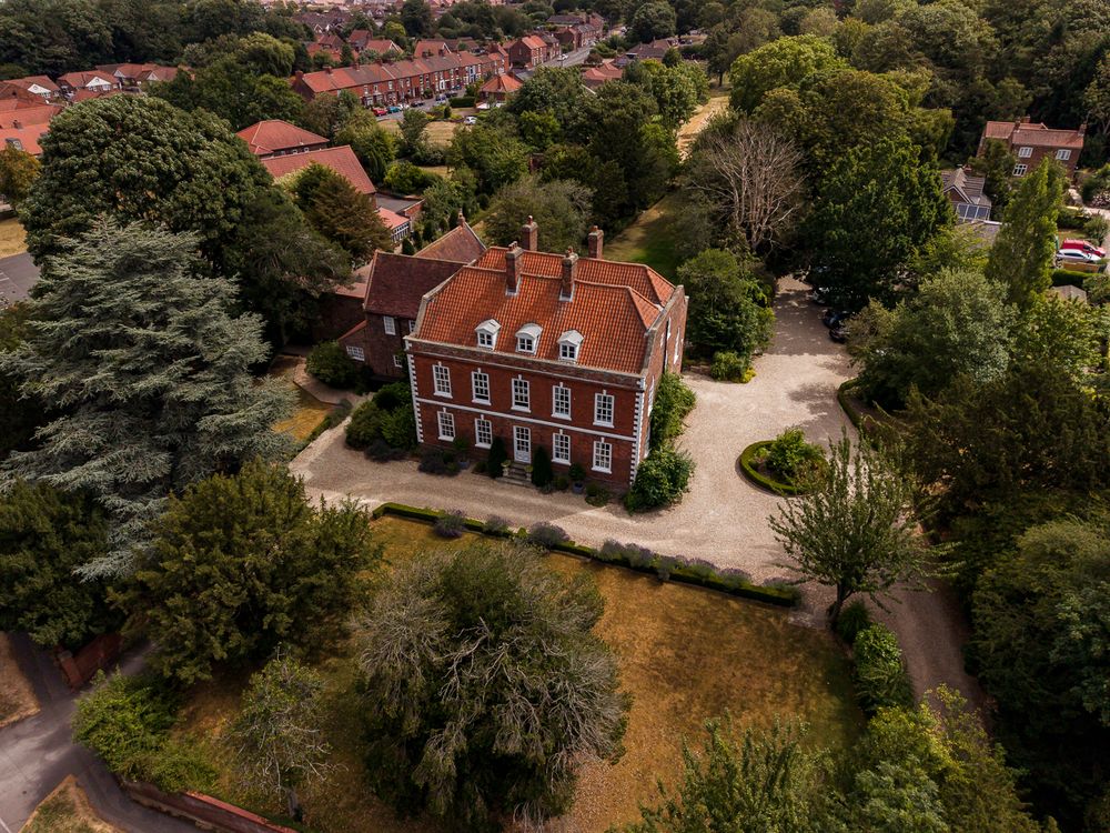 Aerial view of a grand red brick Georgian manor house surrounded by mature trees and landscaped grounds in a suburban setting.