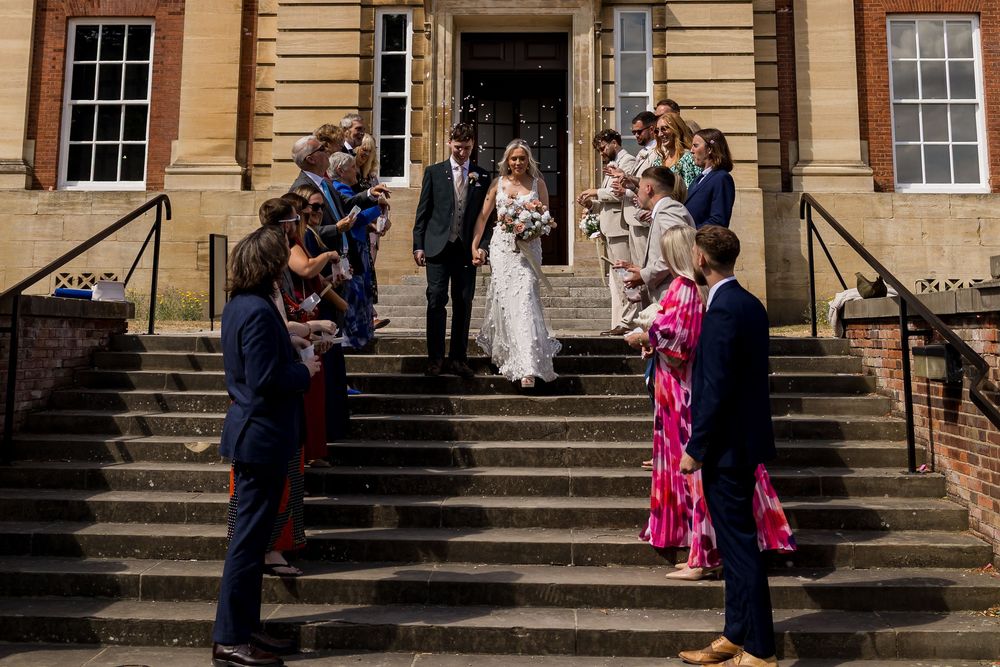 Wedding couple exits elegant building as guests line steps to celebrate with confetti outdoors.