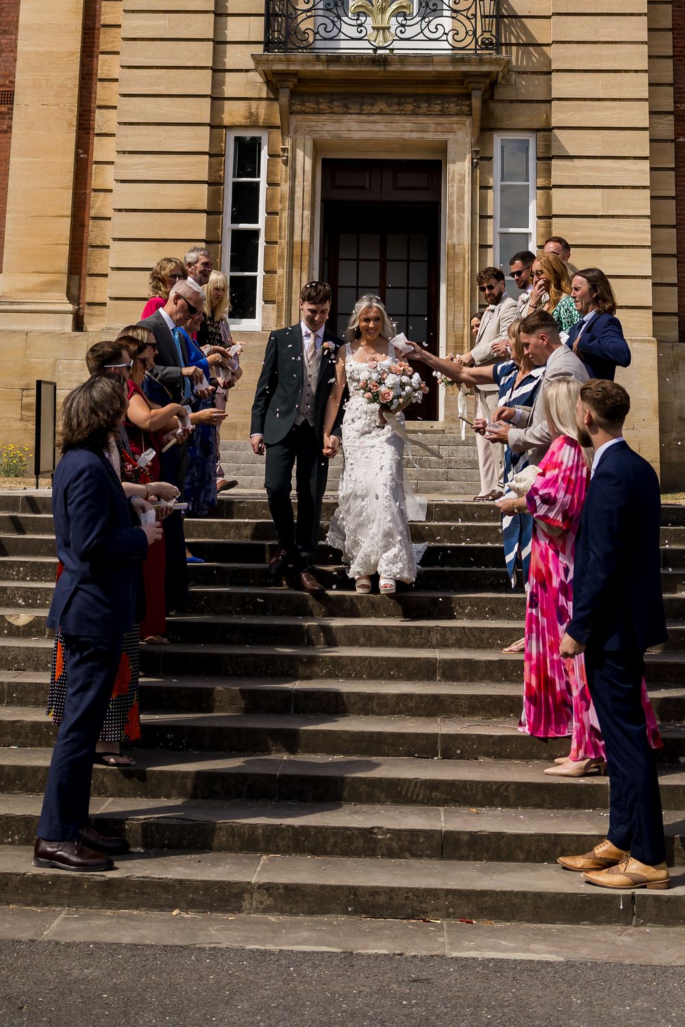 Bride and groom walking down stone steps through confetti arch with wedding guests.