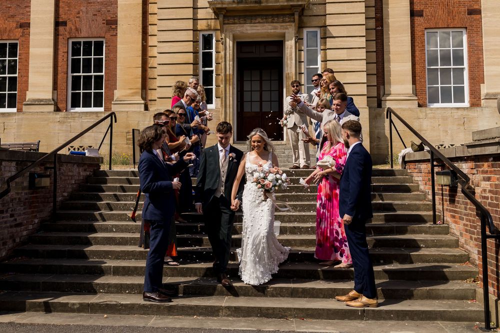 Wedding party celebrating on grand stone steps outside a historic brick building in sunny weather.