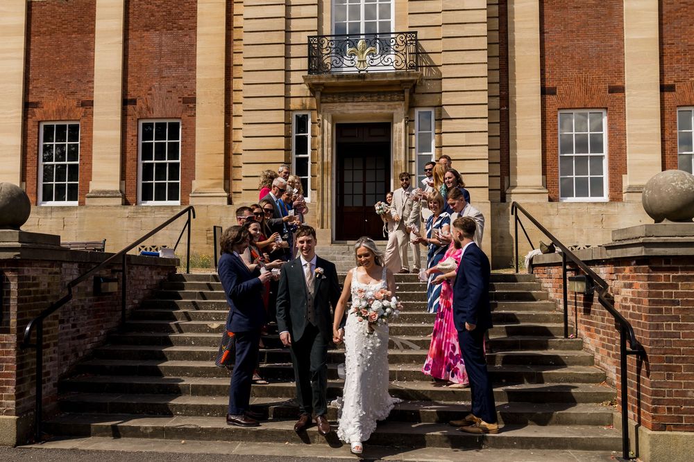 Wedding party descending grand stone steps of historic brick building, bride holding bouquet with guests celebrating.