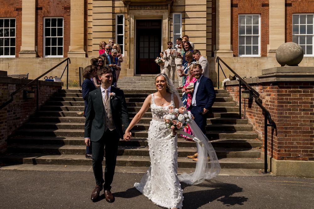 Newlywed couple holds hands and smiles outside a grand historic brick building surrounded by wedding guests.