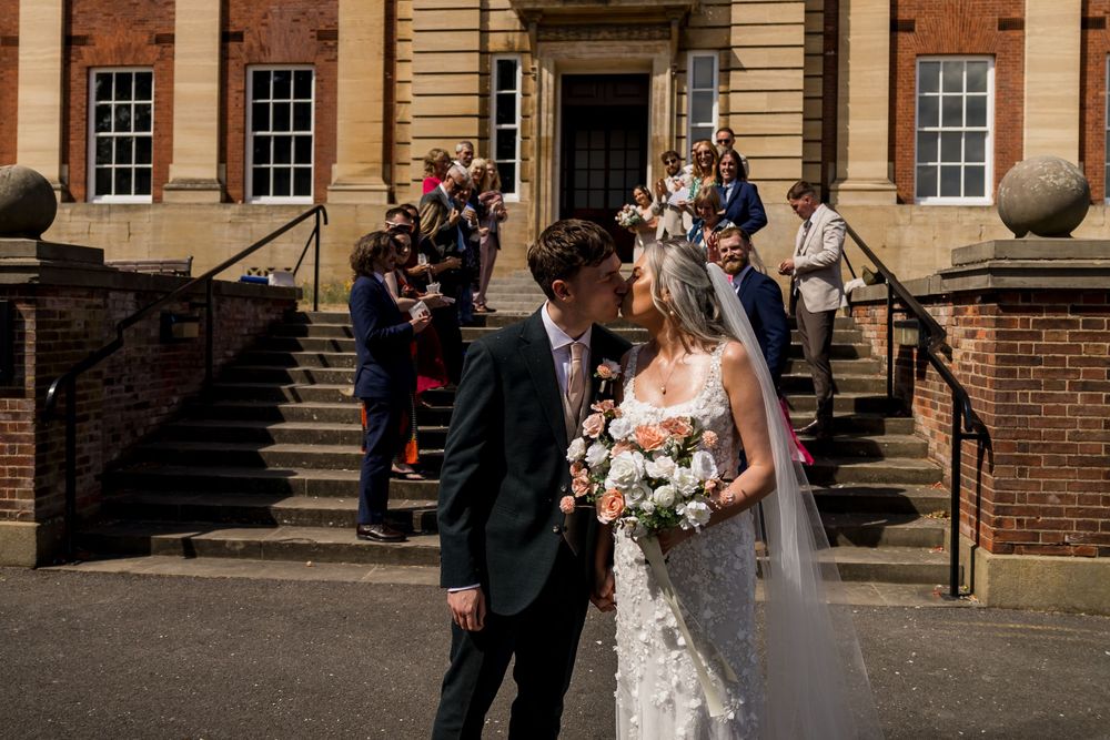 Newlyweds sharing a kiss outside a classic Georgian building, bride in lace gown holding peach floral bouquet.