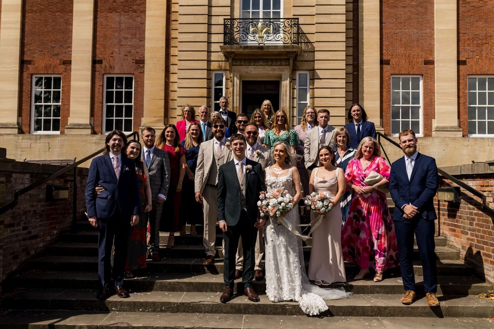 Wedding party group photo on steps of grand brick building on sunny day with bride and groom at center.